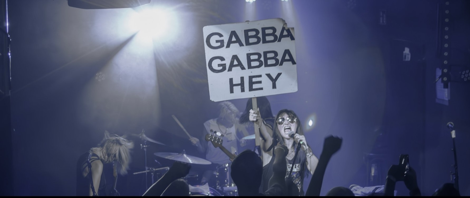 Ein Konzert, man sieht eine Sängerin ein SChild hochhalten, auf dem in großen Lettern "Gabba, Gabba, Hey" steht.