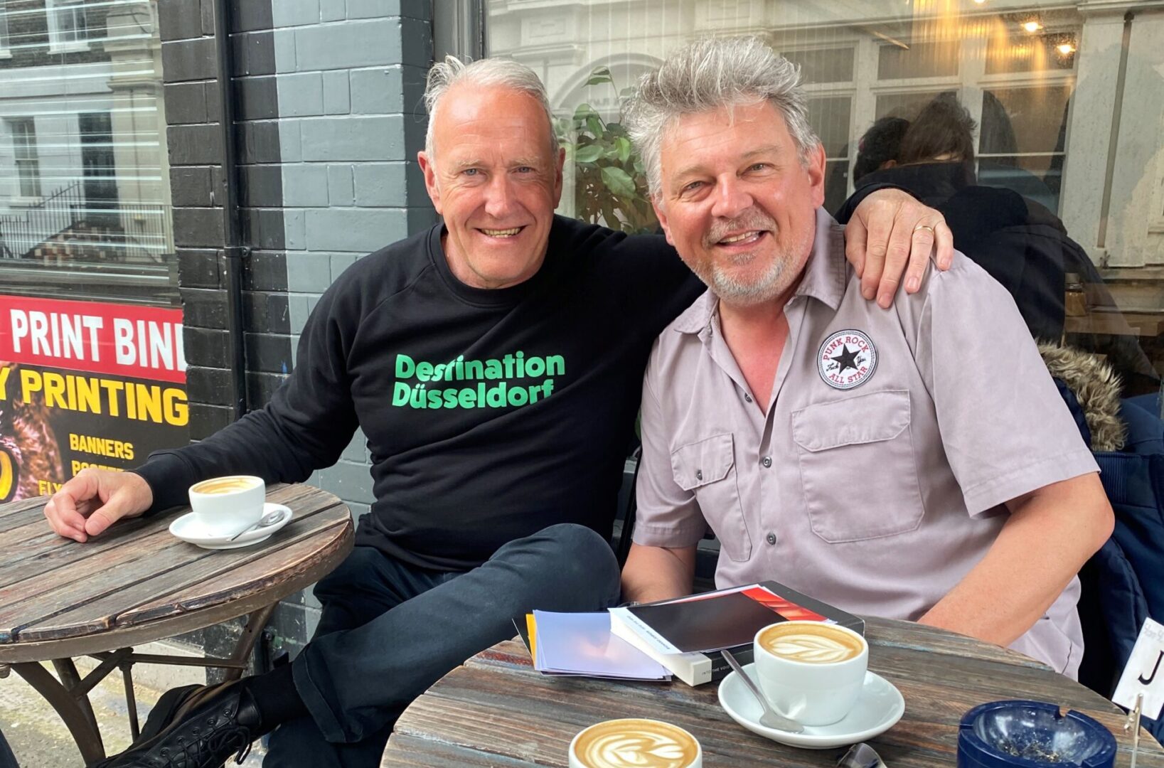 Richard Jobson und Rudi Esch sitzen vor einem Café.