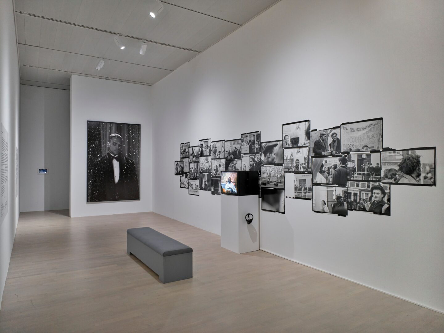 Museumswand mit dicht auf dicht gehängten Schwarzweiß Szenenfotos, davor ein Monitor, auf dem der Film läuft.
