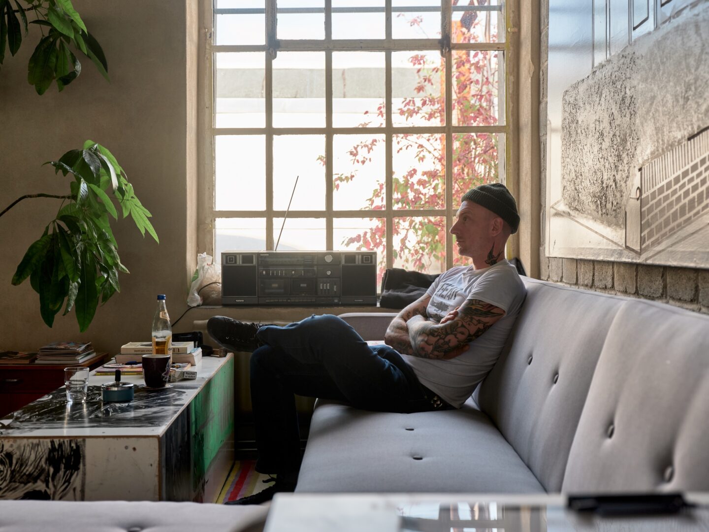 Tattoo Artist Till Pulpanek sitzt auf einem hellgrauen Sofa vor einem alten Fabrikfenster.