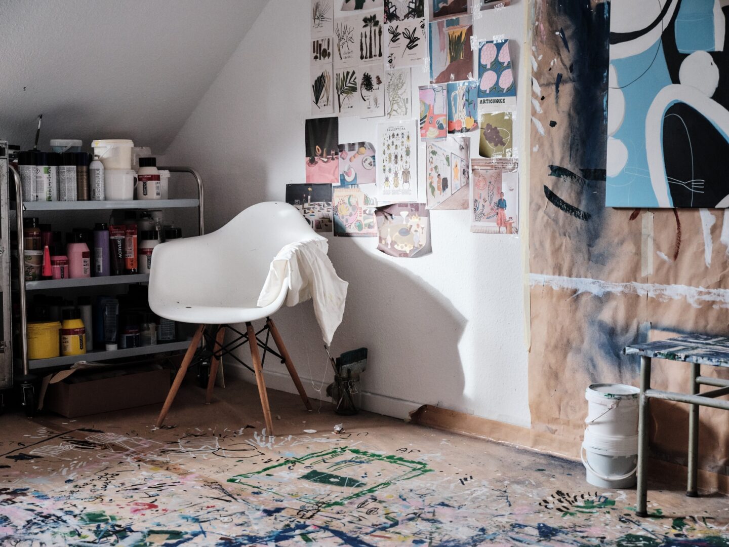 Im Atelier von Anna Pascher, man sieht einen Eames-Plasticarmchair in Weiß in der Ecke vor einem Regal und dem Moodboart, angeschnitten rechts ein Bild in Arbeit.