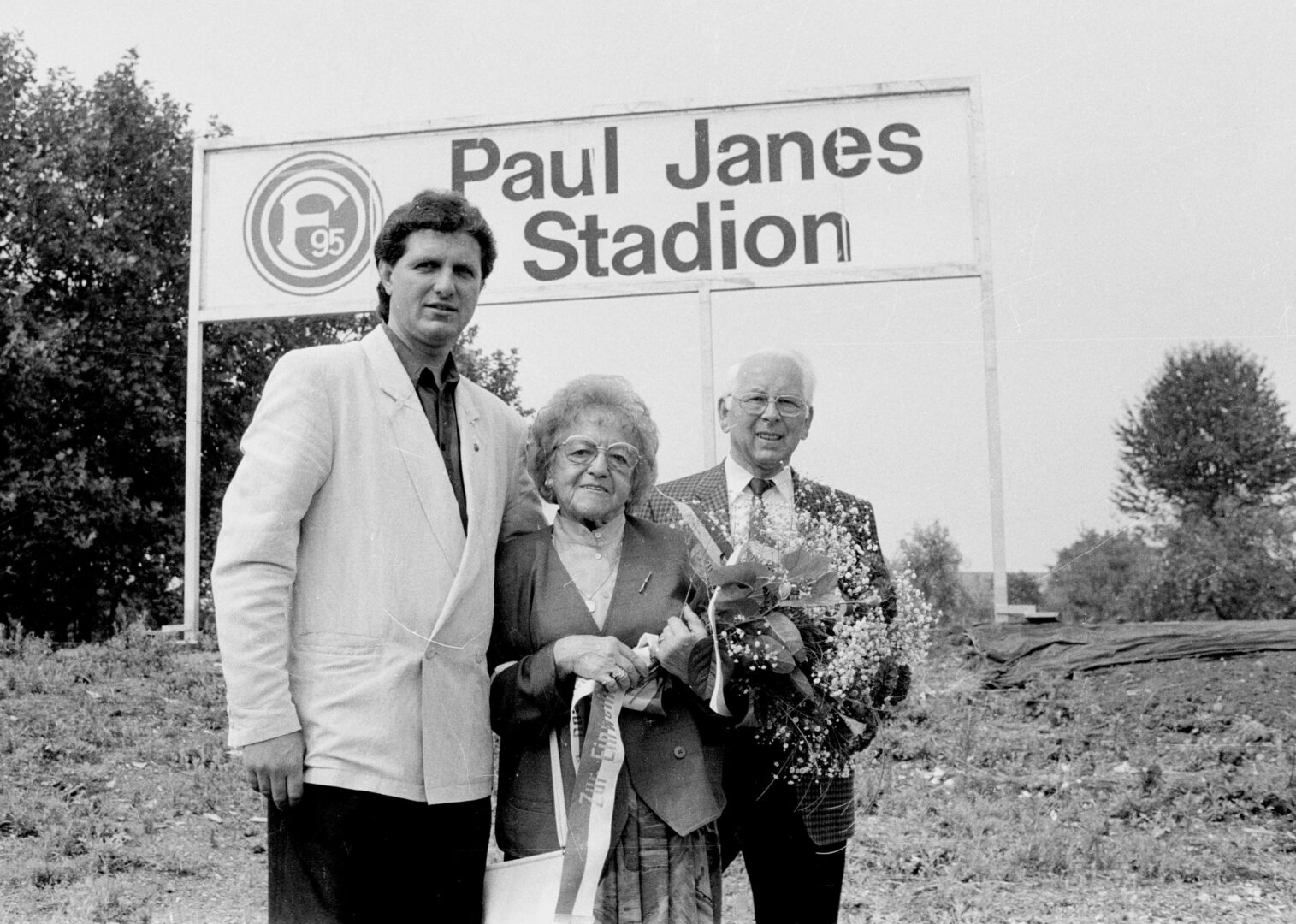 Schwarzweiß Foto: Paul Janes Stadion-Schild und drei Personen stehen davor. 