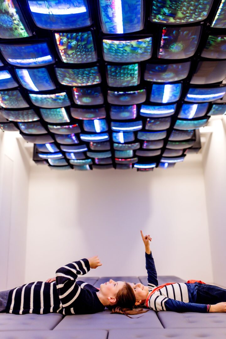 Ein Mädchen und eine Frau in Ringelpullis liegen unter an die Decke montierte Bildschirme auf denen eine Videoinstallation läuft.