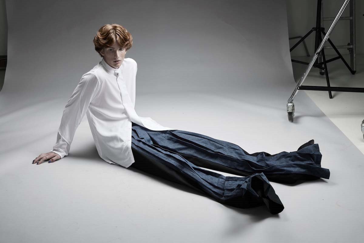 Model im Studio sitzt die Beine weit ausgestreckt in dark blue Hose und weißem Hemd auf dem Boden.