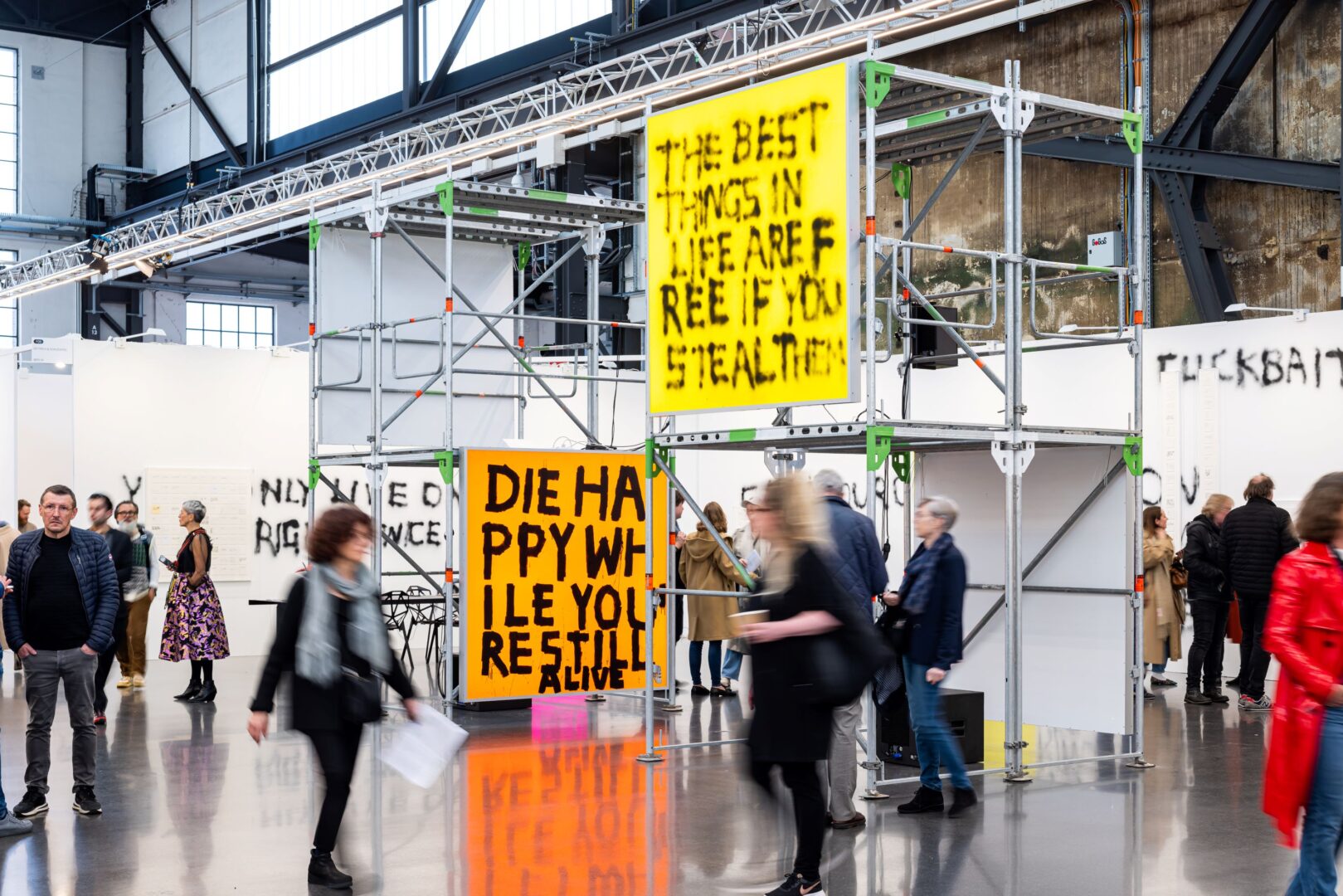 Man sieht zwei beschriftete Banner, die Kunstwerke sind Teil der Art Düsseldorf. Außerdem verwischt und unscharf Besucher*innen der Kunstmesse.