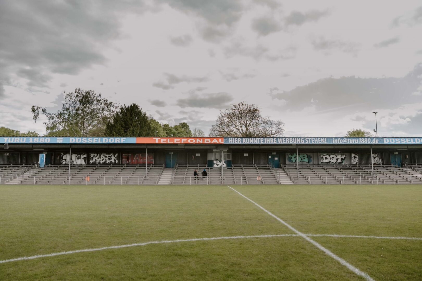 Die Tribüne, vorne das Fußballfeld und oben grauer Wolkenhimmel.