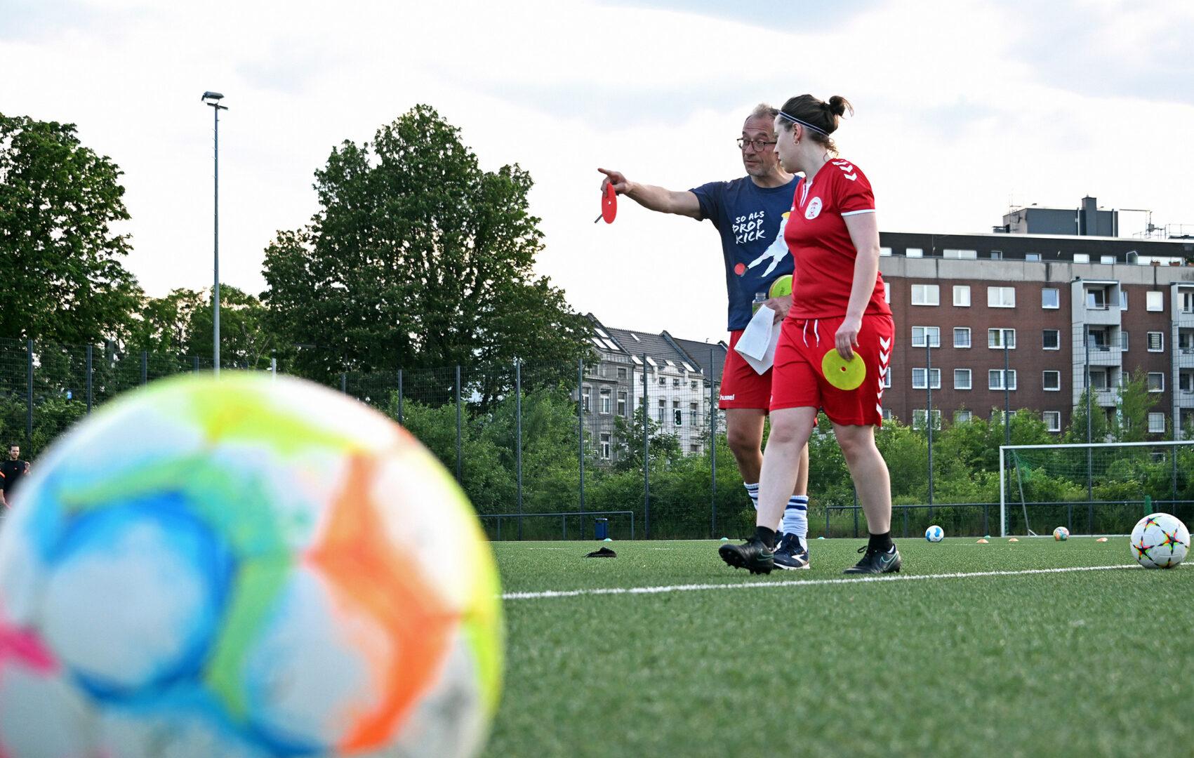 Frauenfußball: Im Vordergrund ein Fußball auf dem Rasen, im Hintergrund der Trainer mit einer Fußballspielerin.