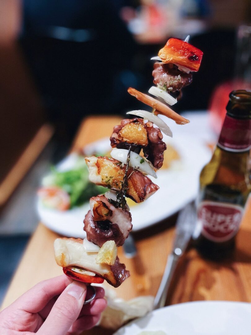 Man sieht einen Oktopus-Spieß vom Grill, gehalten von einer Hand, im Hintergrund einen Flasche Super Bock Bier.