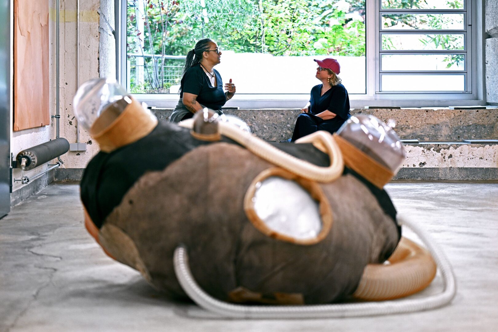 Vorne eine Skulptur, hinten sitzen Anys Reimann (links) und Karolina Landowski vor einem Panoramafenster und unterhalten sich.