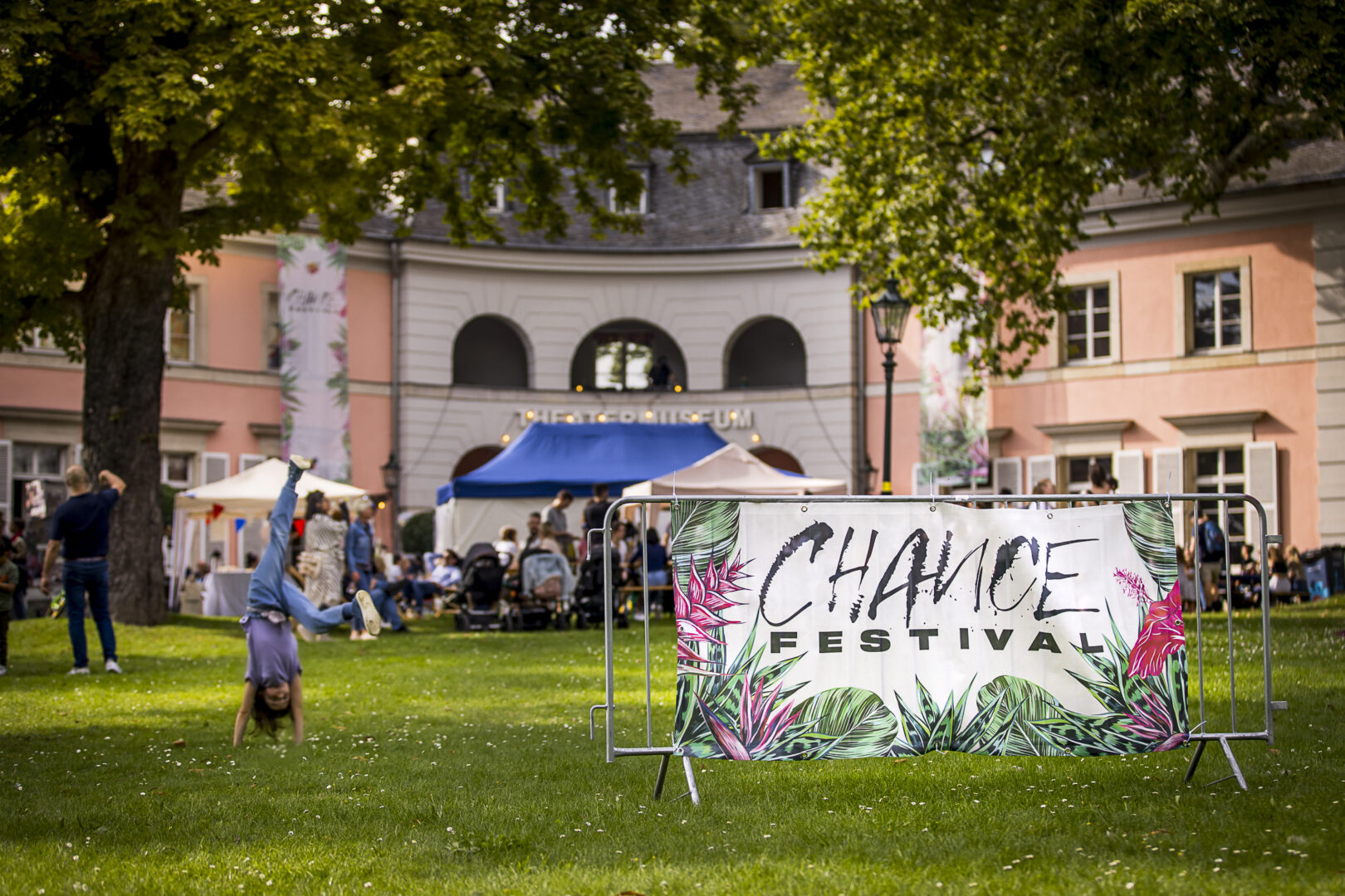 Ein Banner, auf dem steht: Chance Festival ist auf einer grünen Wiese im Hintergrund sieht man das Theatermuseum und Menschen.