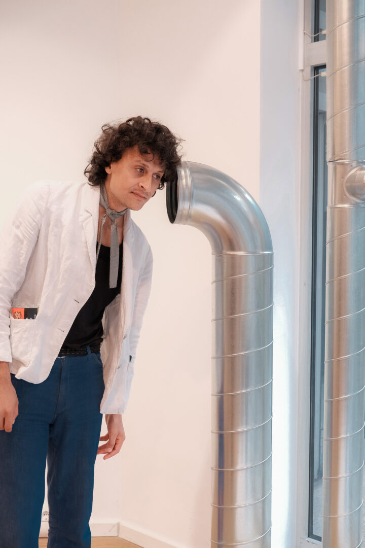 Mann in weißem Sakko, hält sein Ohr an ein silbriges "Ofenrohr", das Teil einer Installation ist.