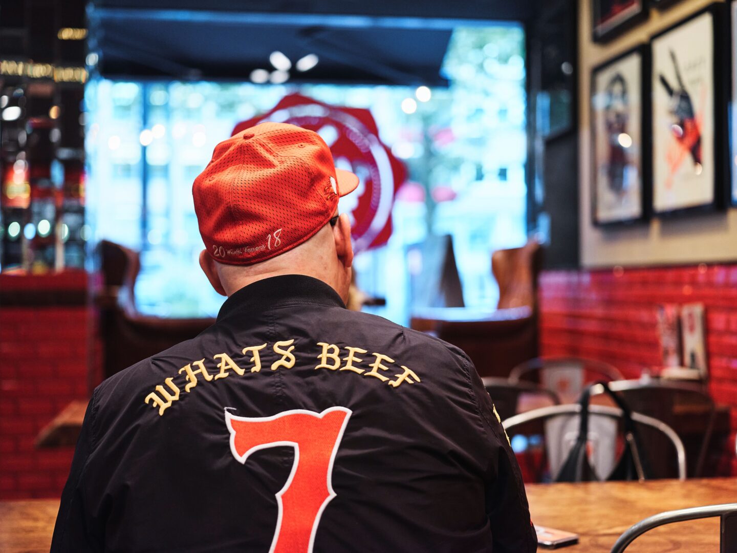 Selim am Tisch sitzend von hinten fotografiert. Er trägt eine Baseball-Jacke, auf der What's Beef und darunter eine 7 gestickt ist.