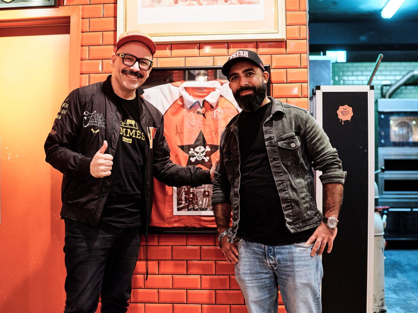 Selim (re) und sein Partner Ali stehen vor einer rot gekachelten Wand im Burgerladen.