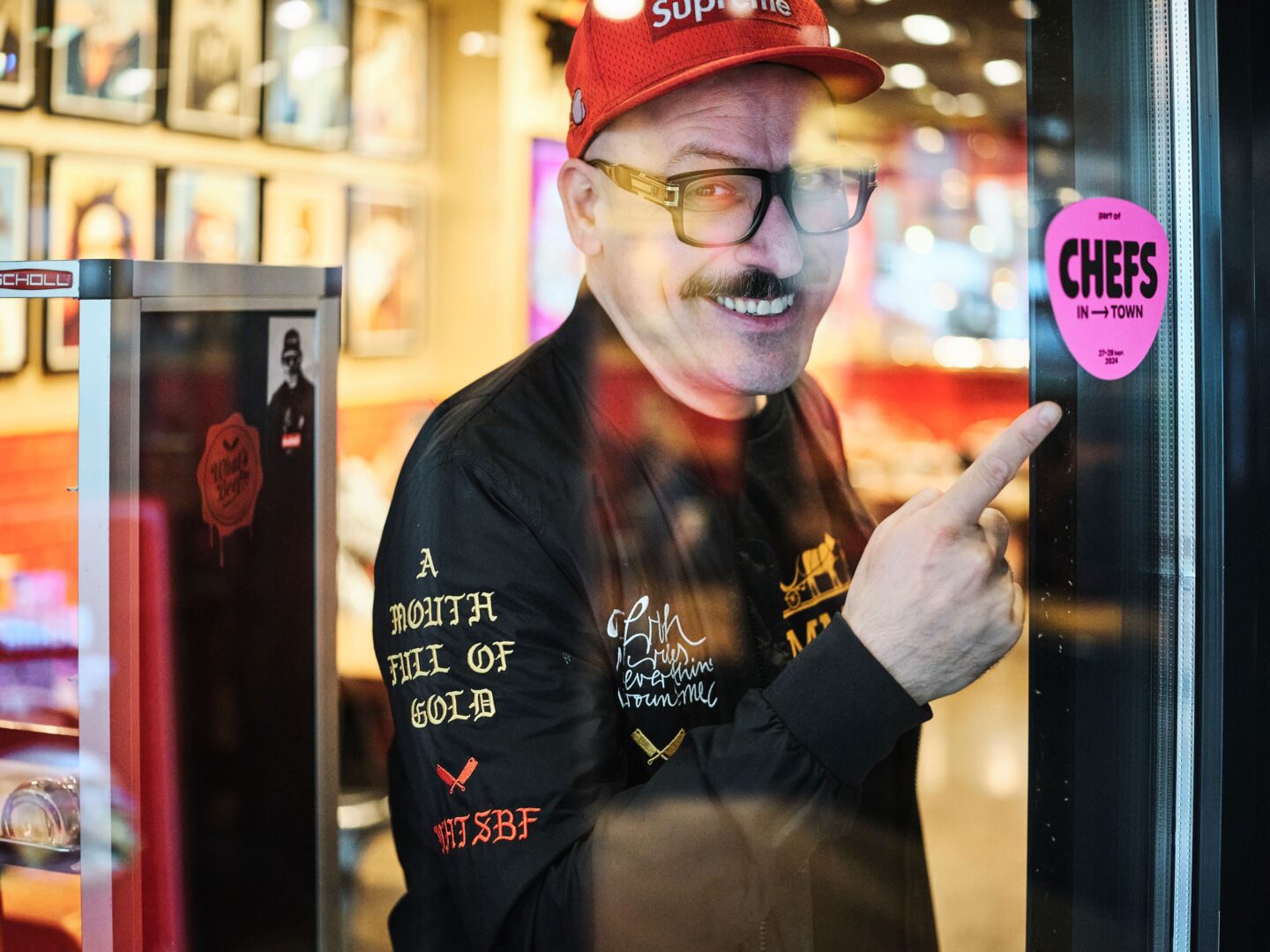 Selim von außen durch die Schaufensterscheibe fotografiert, wie er auf den pinken "Chefs in Town"-Aufkleber zeigt.