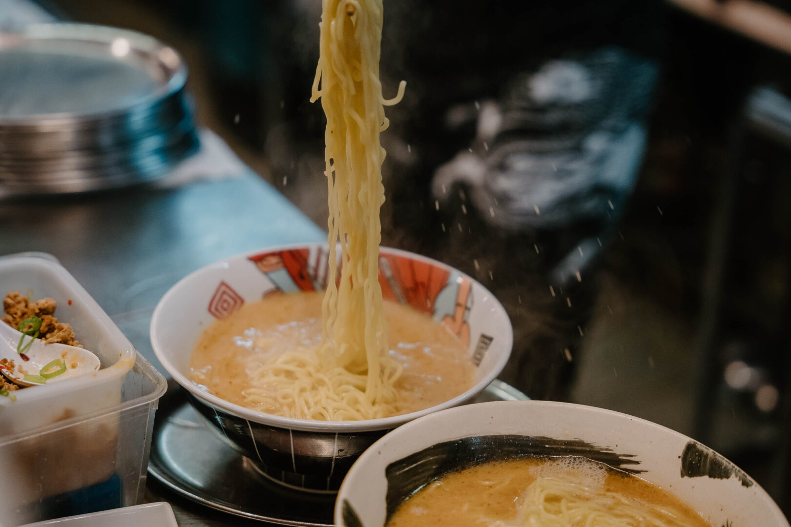 Man sieht zwei Ramen-Schüsseln, in eine werden die Ramennudeln gerade serviert, damit sie an den Tisch gebracht werden können.