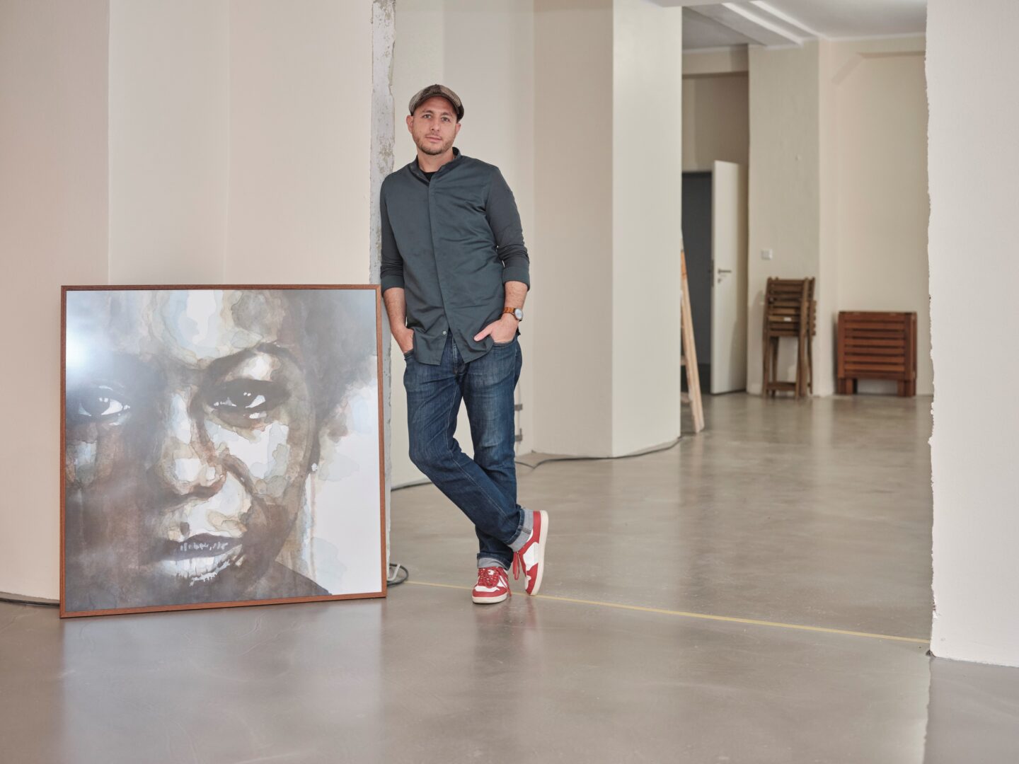 Sofian Neubauer lehnt an eine weiße Wand, an der links ein Bild steht, man sieht das Portrait einer schwarzen Frau. Im Hintergrund sieht man leere Räume.