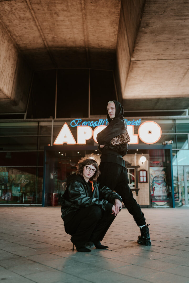 Maurice steht und Daria kniet vor dem Eingang des Apollo-Theaters am Rhein.