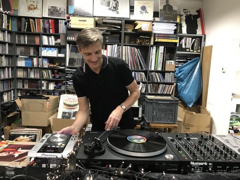 Marc Meyer in einem Polohemd legt Platten auf. Im Hintergrund sieht man Regale voll von CDs und Vinylplatten.