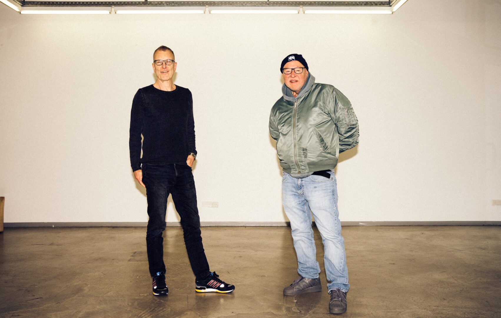 Hubert Ostendorf (li) und Mike Litt stehen in der leeren Galerie und schauen in die Kamera.