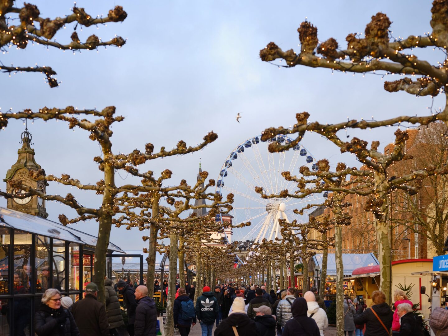Man sieht die Promenade, rechts und links die mit Lichterketten geschmückten Bäume und hinten das Riesenrad. Der Himmel ist noch blau, kurz bevor die Sonne untergeht.