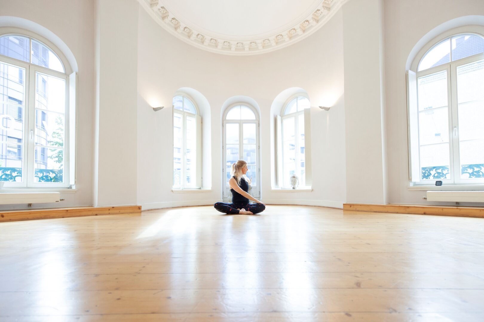Ein sehr großer, hoher und leerer Raum mit hellem Holzboden, hinten sind mehrere Bogenfenster. In der Mitte sitzt eine Frau und macht Yoga im Lotussitz.