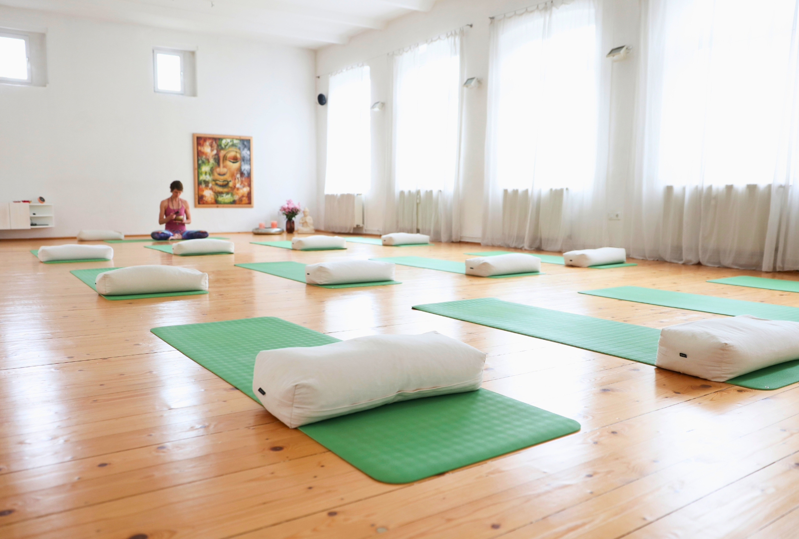 Yogastudio mit Holzboden, grünen Matten und man sieht im Hintergrund die Yogalehrerin.