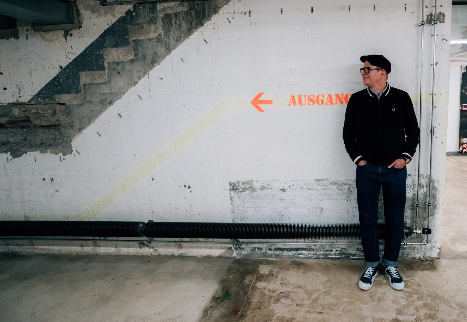 Tobi Dahmen vor einer weiß gestrichenen Backsteinwand. Er schaut nach links. In Schulterhöhe hinter ihm der Schriftzug Ausgang in knallorange.