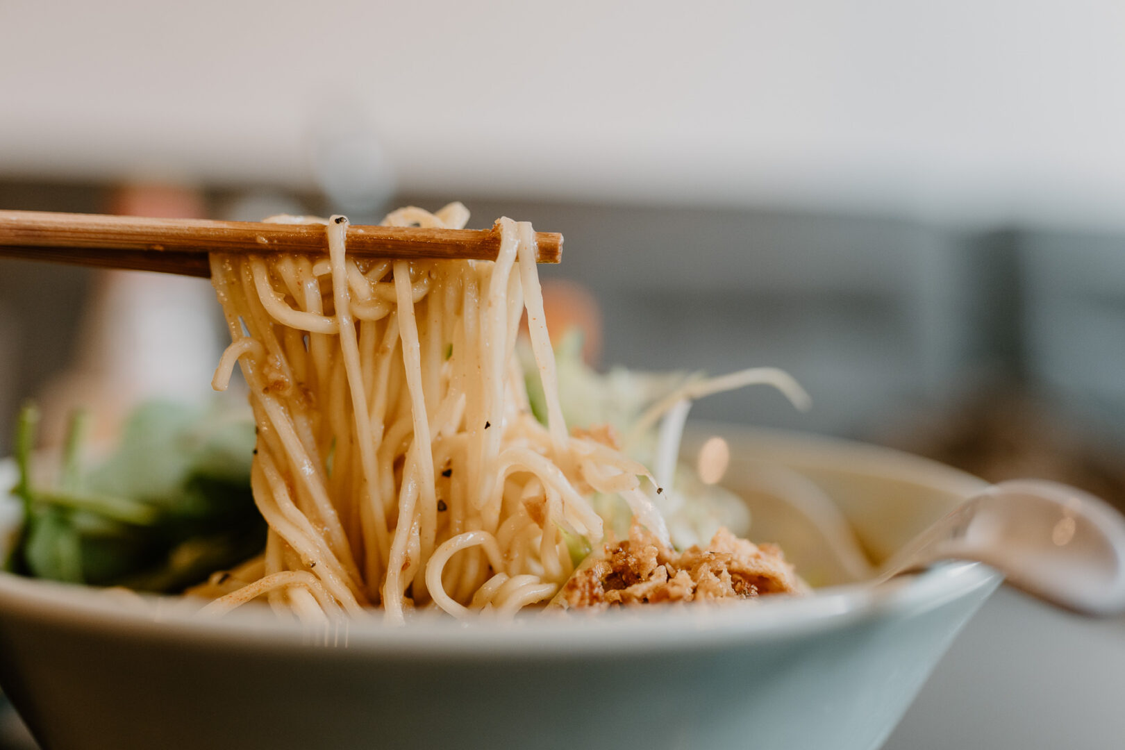 Nahaufnahme einer Schüssel Ramen von der Seite. Man sieht zwei Stäbchen, die gerade eine Portion Nudeln aus der Schüssel heben.