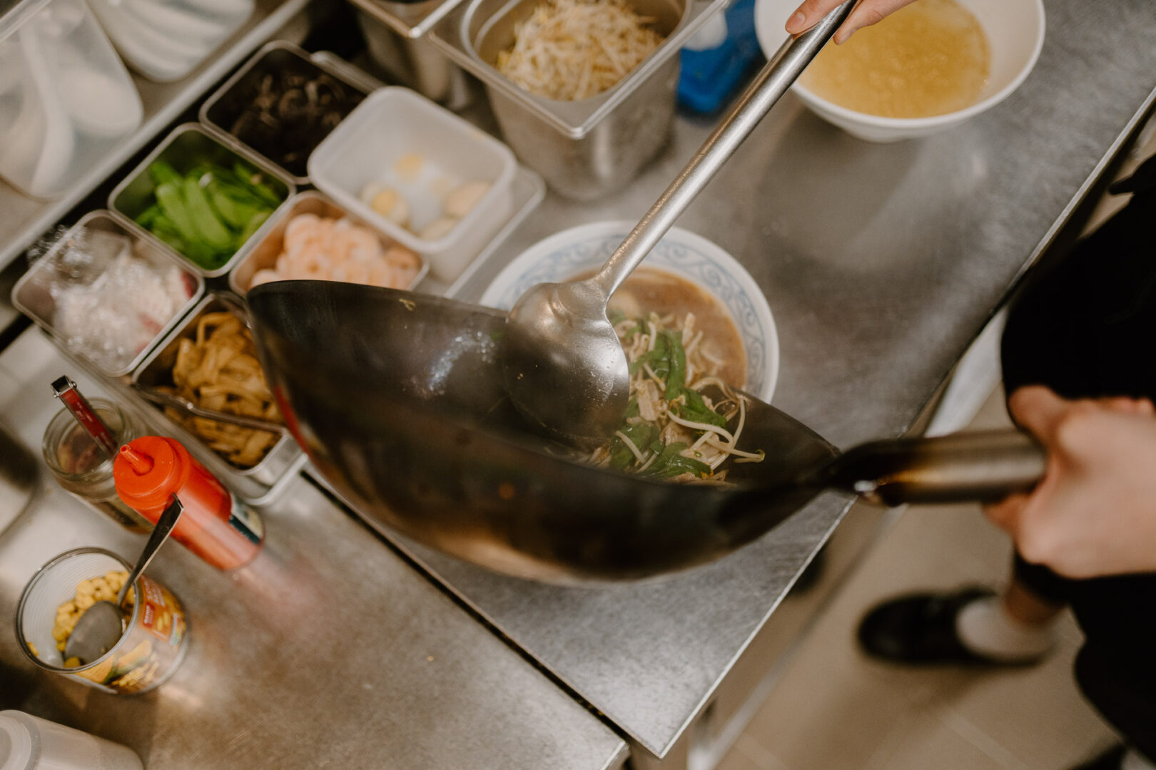Man blickt sozusagen dem Ramen-Koch über die Schulter, wie er Brühe in eine angerichtete Schüssel Ramen gießt.