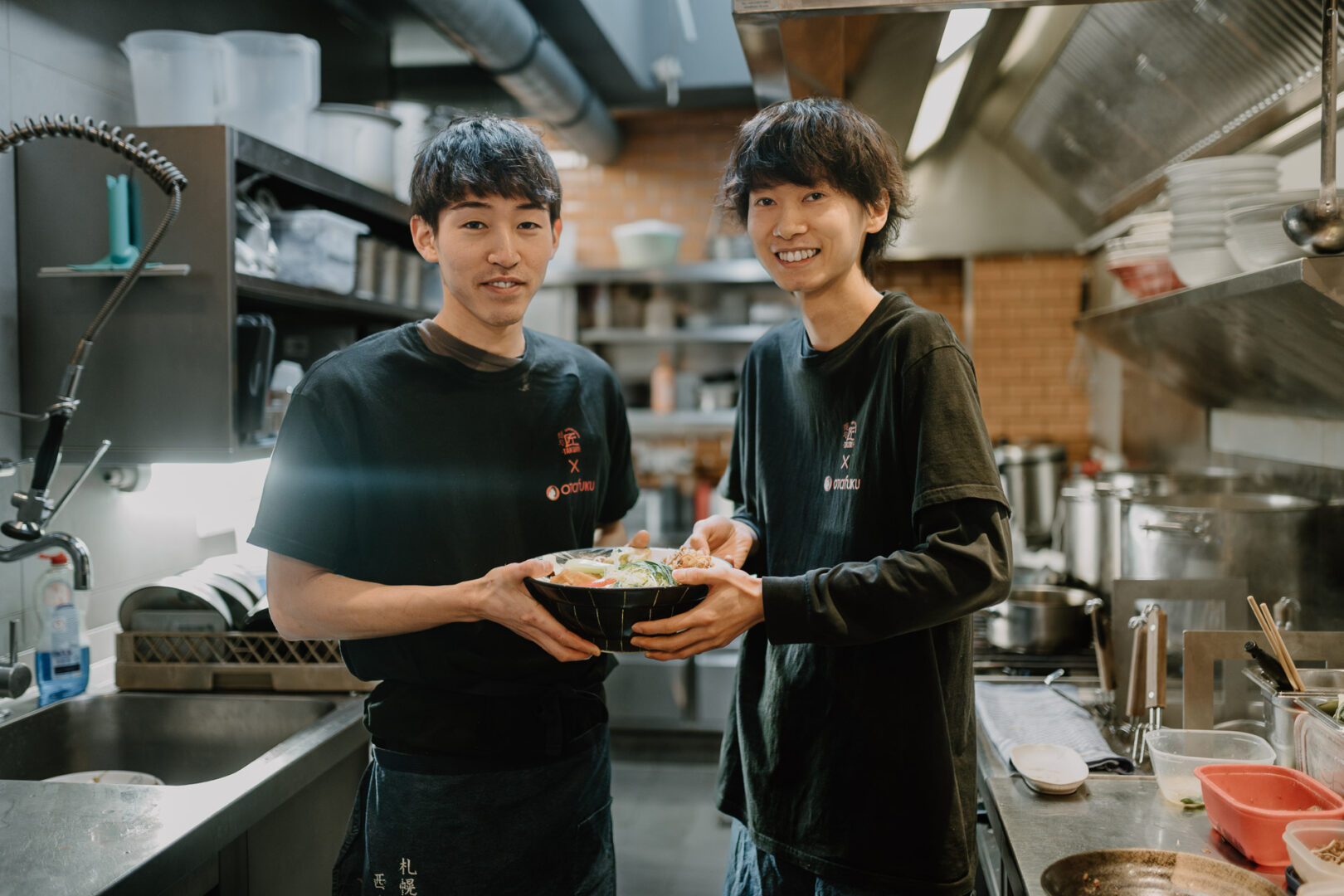 Man sieht zwei japanische Männer, die zwischen sich eine Schüssel Ramen halten und in der Küche von Takumi Veggie & Chicken stehen.