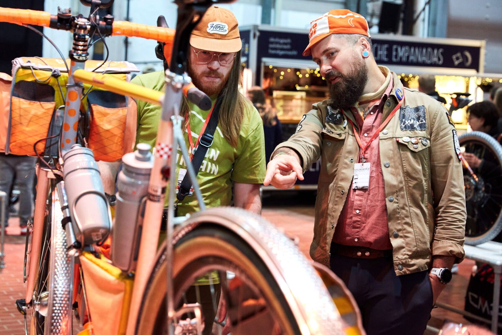 Zwei bärtige Männer mit orangefarbenen Kappen sprechen über ein Fahrrad, das im Anschnitt vorne links zu sehen ist. 