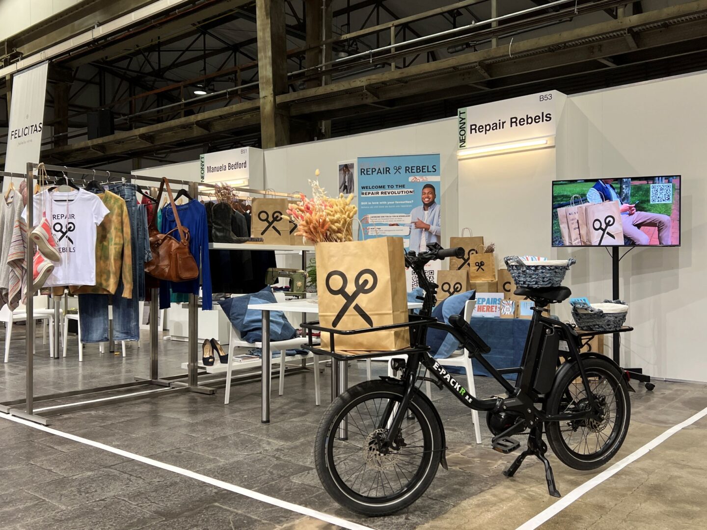 Ein Messestand von Repair Rebels. Man sieht im Vordergrund ein schwarzes Fahrrad und im Hintergrund Displays und eine Kleiderstange mit Kleidungsstücken. 