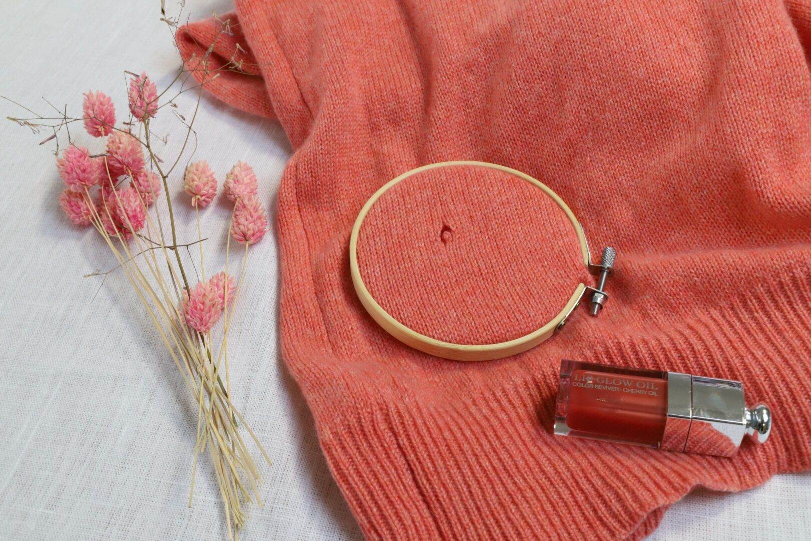 Man sieht einen orangeroten Pulli mit einem kleinen Loch, das in einen Rahmen zur Reparatur eingespannt ist. Links liegen farblich passende Blumen.