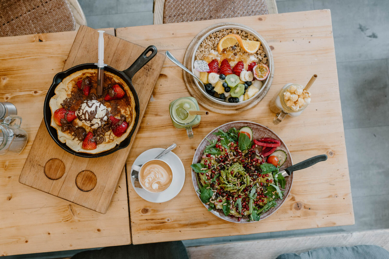 Man sieht einen Holztisch von oben, darauf angerichtete Frühstücksspeisen in einer gusseisernen Pfanne ein Pfannekuchen mit Erdbeeren, Schoko & Eis; auf einem Teller SAlat mit Granatäpfeln und ein Müsli. 