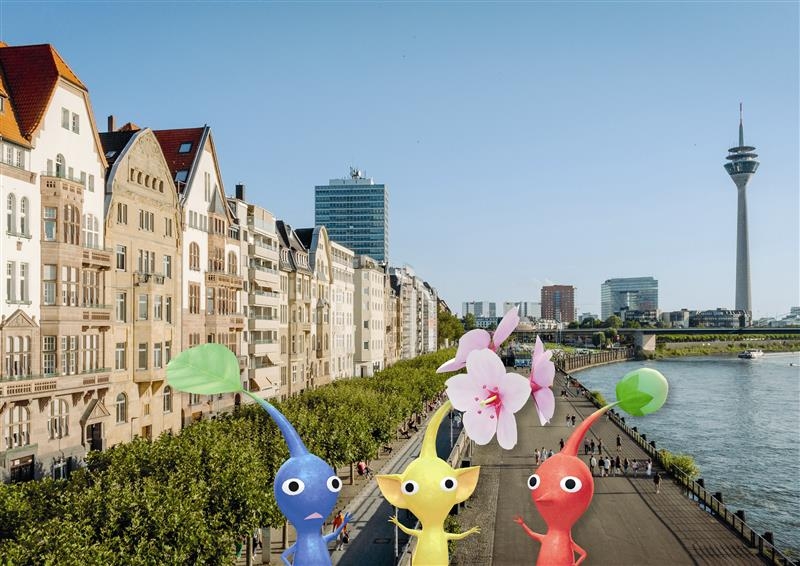 Man sieht drei Pikmins, Figuren von Nintendo, vor einem echten Foto der Rheinuferpromenade. 