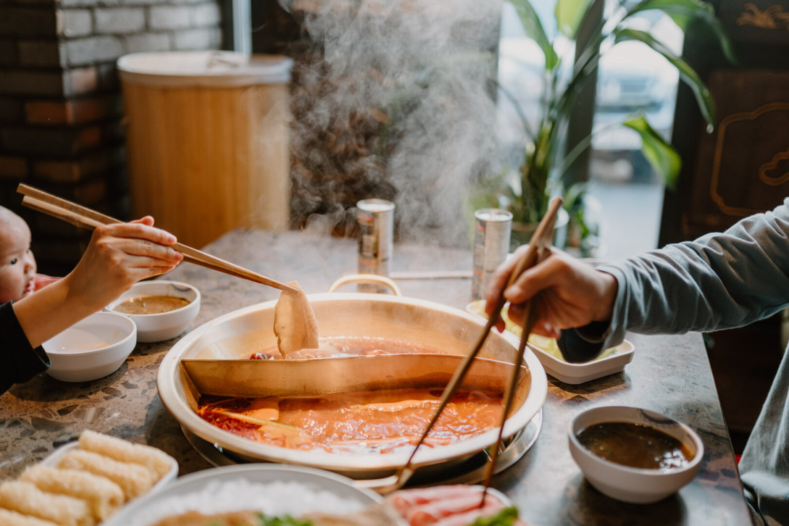 Man sieht einen Hot Pot mit viel Brühe und Hände mit Stäbchen, die Zutaten in die Brühe tunken.