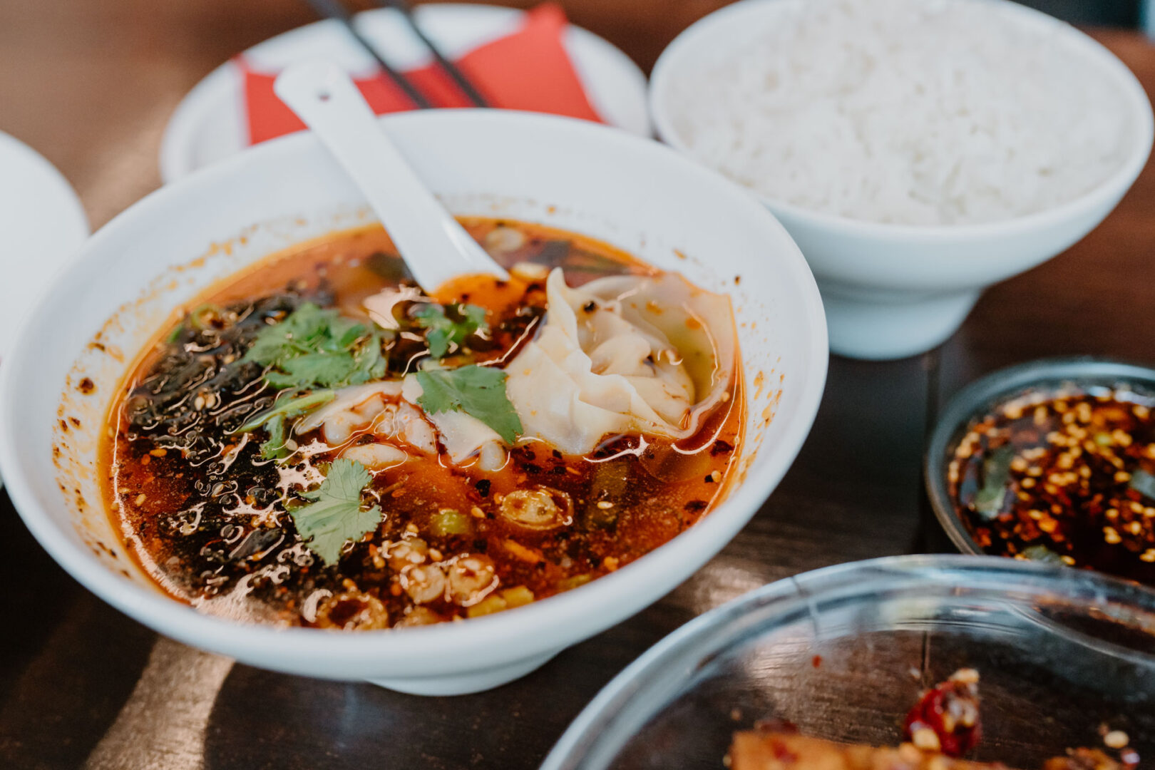 Man sieht im Mittelpunkt eine weiße Schale mit einer Brühe, die sehr rot von Chili-Öl ist und ein paar Dumplings.