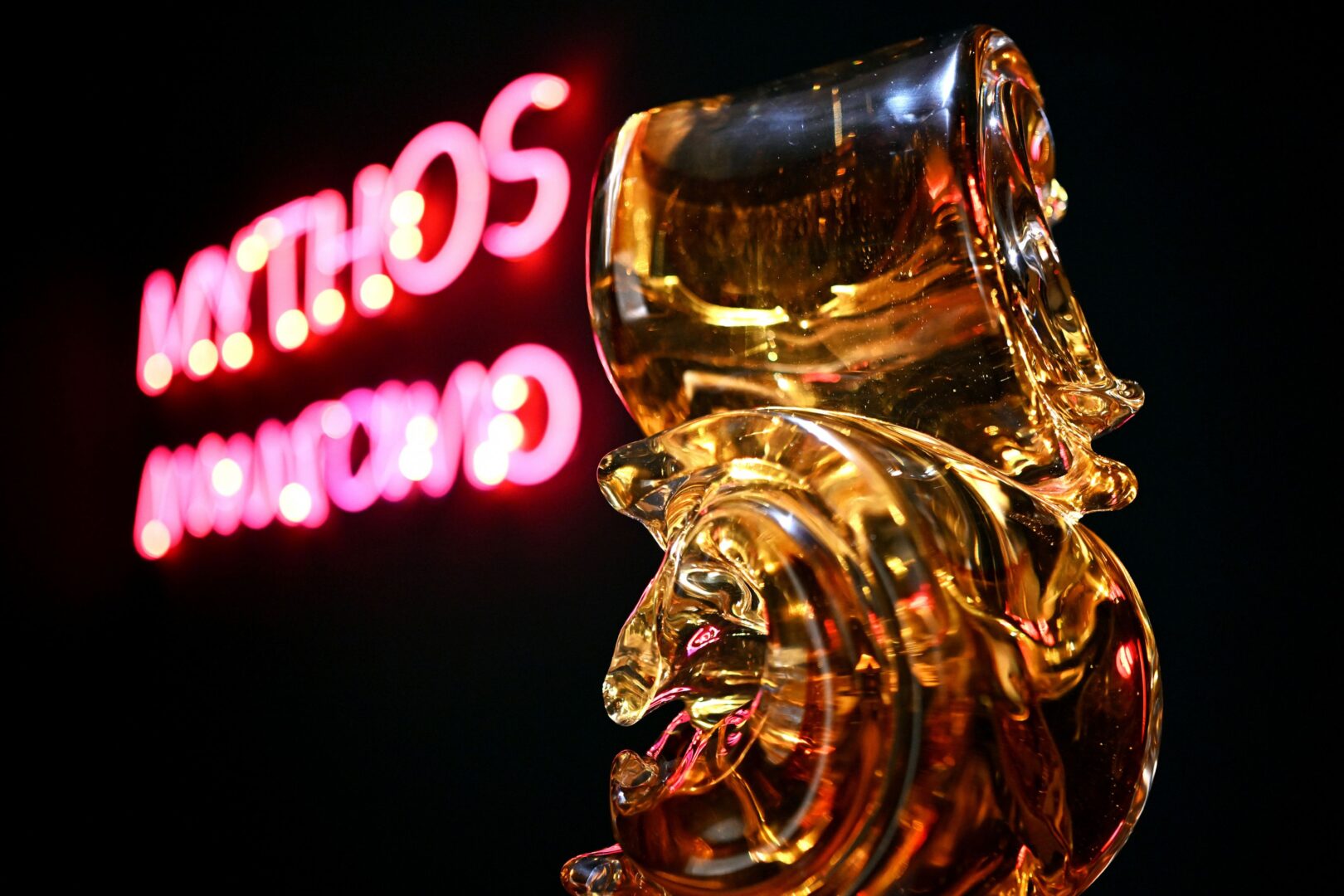 Man sieht eine bernsteinfarbene Glasskulptur. Im Hintergrund leuchtet unscharf der Neonschriftzug Mythos Murano.