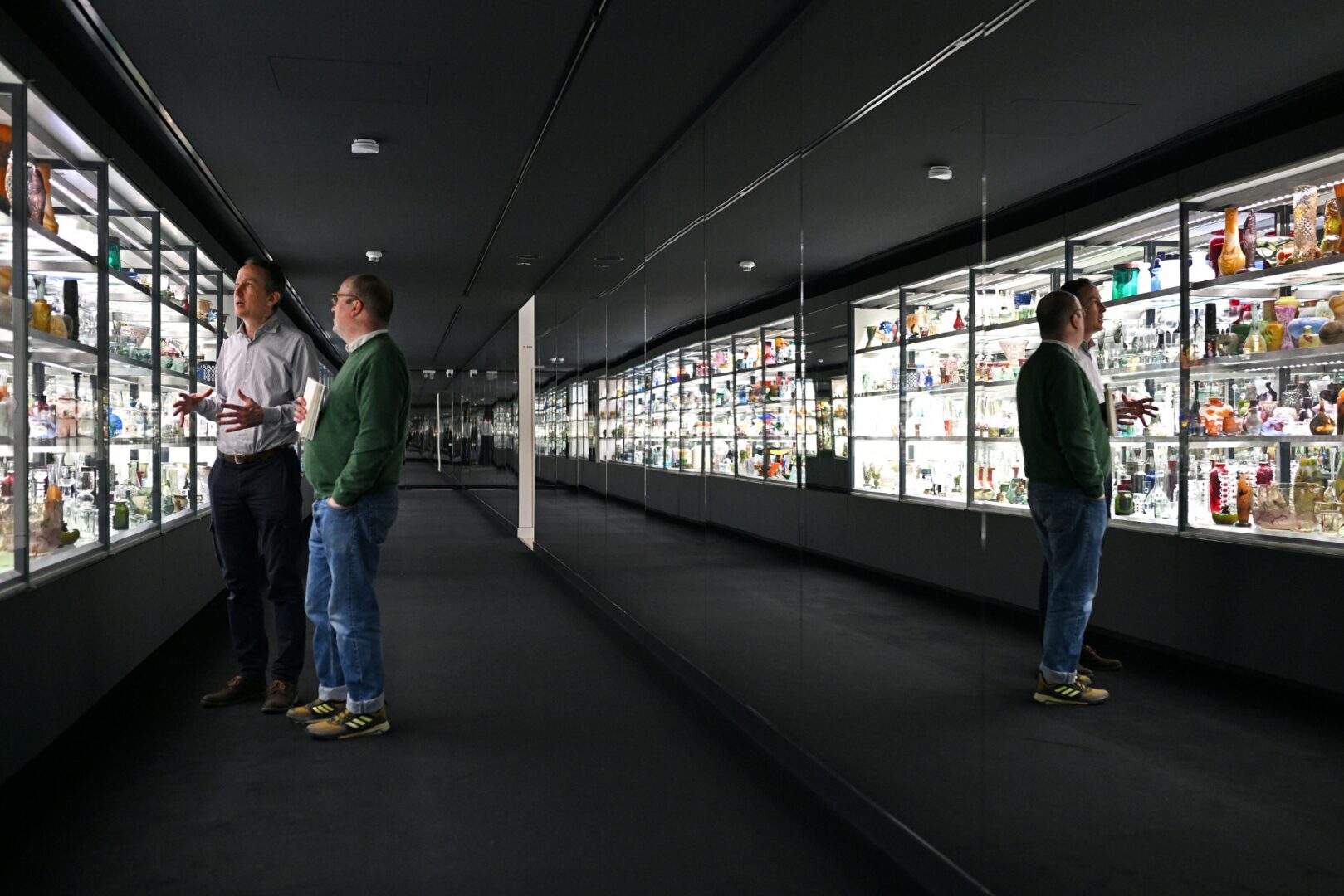 Man sieht das Spiegelkabinett der Glassammlung. Rechts sind die Regale voller gläserner Ausstellungsstücke, links werden sie gespiegelt, so dass in dem dunklen Raum eine Art Gasse zu entstehen scheint, flankiert von beleuchteten Regalen rechts und links.