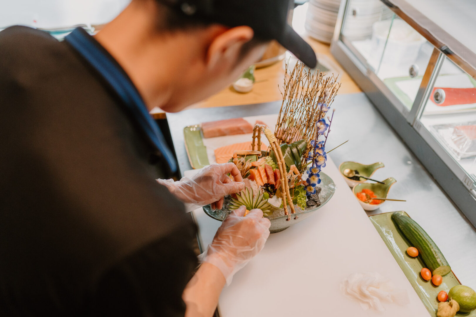Blick über die Schulter des Kochs, der einen sehr bezaubernd angerichteten Teller mit Lachs-Sashimi eine dünn gefächerte Gurke drapiert.