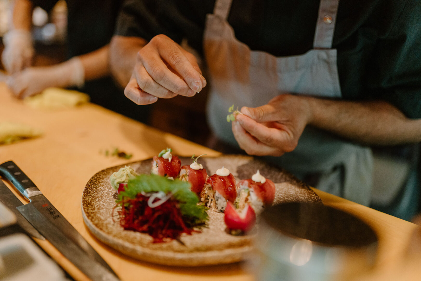 Man sieht die Hände eines Kochs, der etwa Kresse behutsam auf die angerichteten Sushi-Rollen gibt.