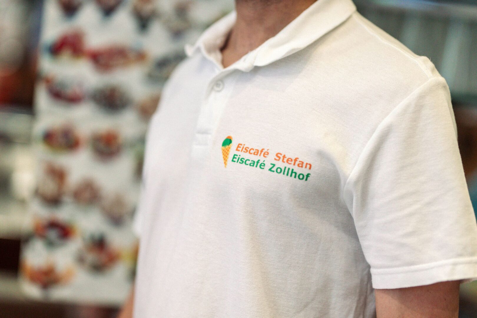 Man sieht ein Polohemd, getragen von einem Kellner. Man sieht nur das weiße Polohemd und den Aufdruck: Eiscafé Stefan, darunter Eiscafé Zollhof.