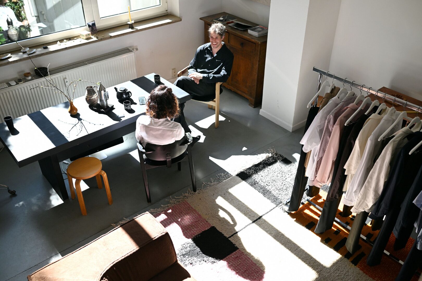 Von oben fotografiert, sieht man die Autorin und den Cornelius an einem langen Holztisch sitzen. Licht fällt durch ein großes Fenster und wirft Schatten auf den Boden. 
