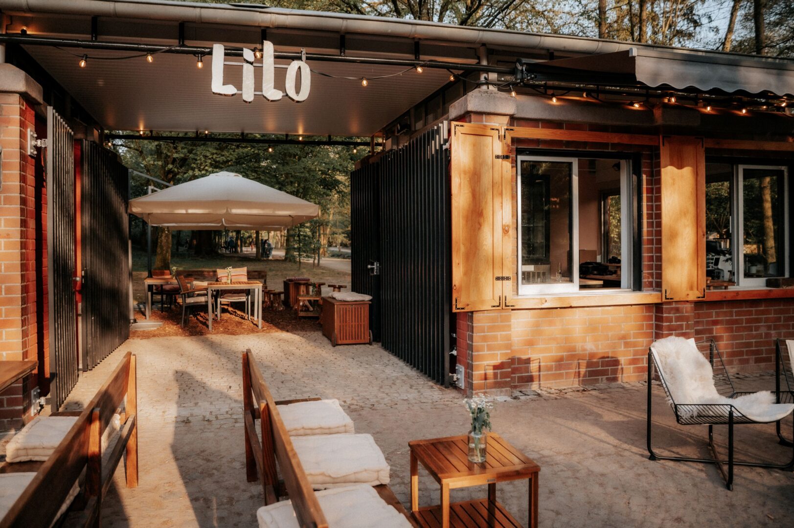 Man sieht den Eingang und die vordere Terrasse des Cafés Lilo. Bänke, Tische und auch das Café sind aus Holz. Es sieht sehr rustikal aus.