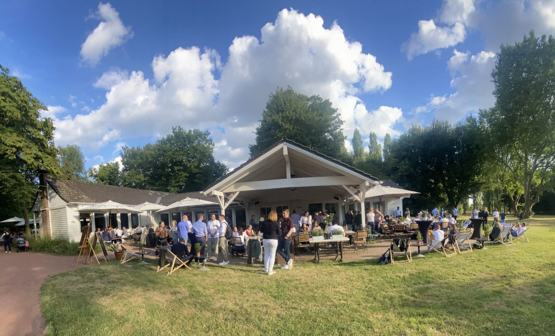 Man sieht eine Weitwinkelaufnahme der Westterrasse des Nordpark Cafés. Über dem Dach des Cafés türmen sich weiße Wolken gegen einen strahlend blauen Himmel.