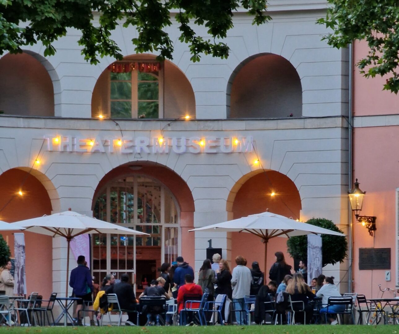 Im Hintergrund ein Ausschnitt des Theatermuseum im Hofgarten. Vom Balkon hängt eine Lichterkette. Darunter sieht man auf der Wiese Tische, Sonnenschirme und Menschen. Es ist bereits Abend und etwas dunkler.