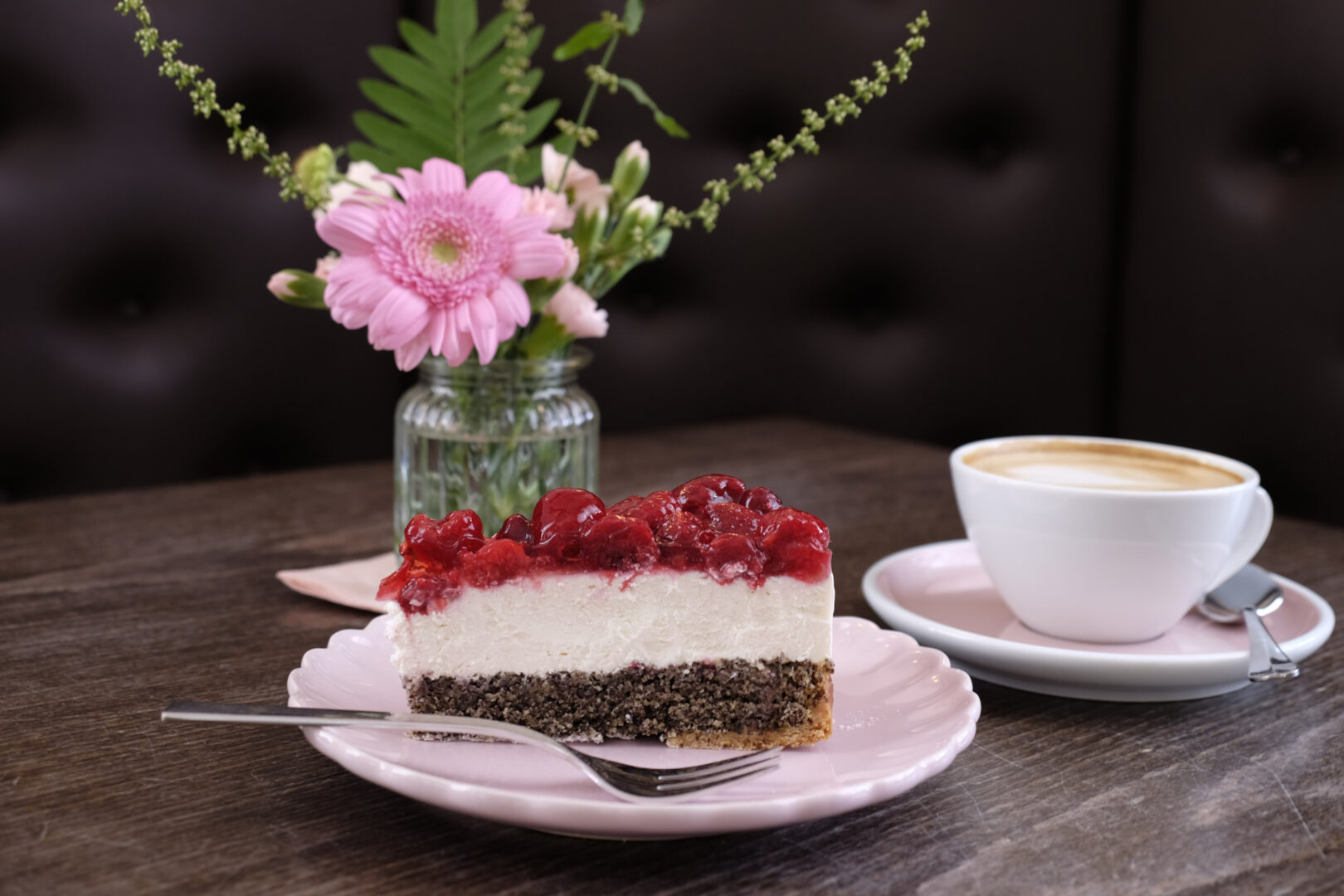 Man sieht einen schön eingedeckten Tisch mit einem Stück Torte, einer Tasse Kaffee und einer pinken Gerbera in einer Vase.