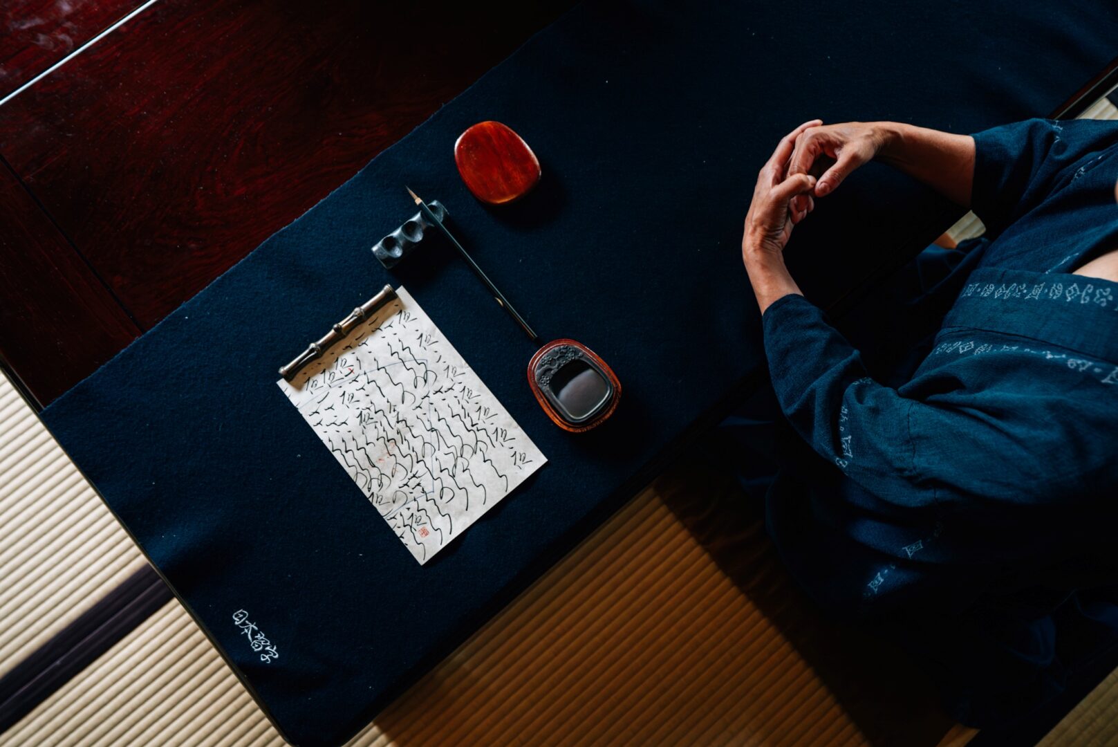 Von oben fotografiert, sieht man einen Tisch, darauf Ayas gefaltete Hände und links davon ein Papier mit kalligrafischen Schreibübungen.