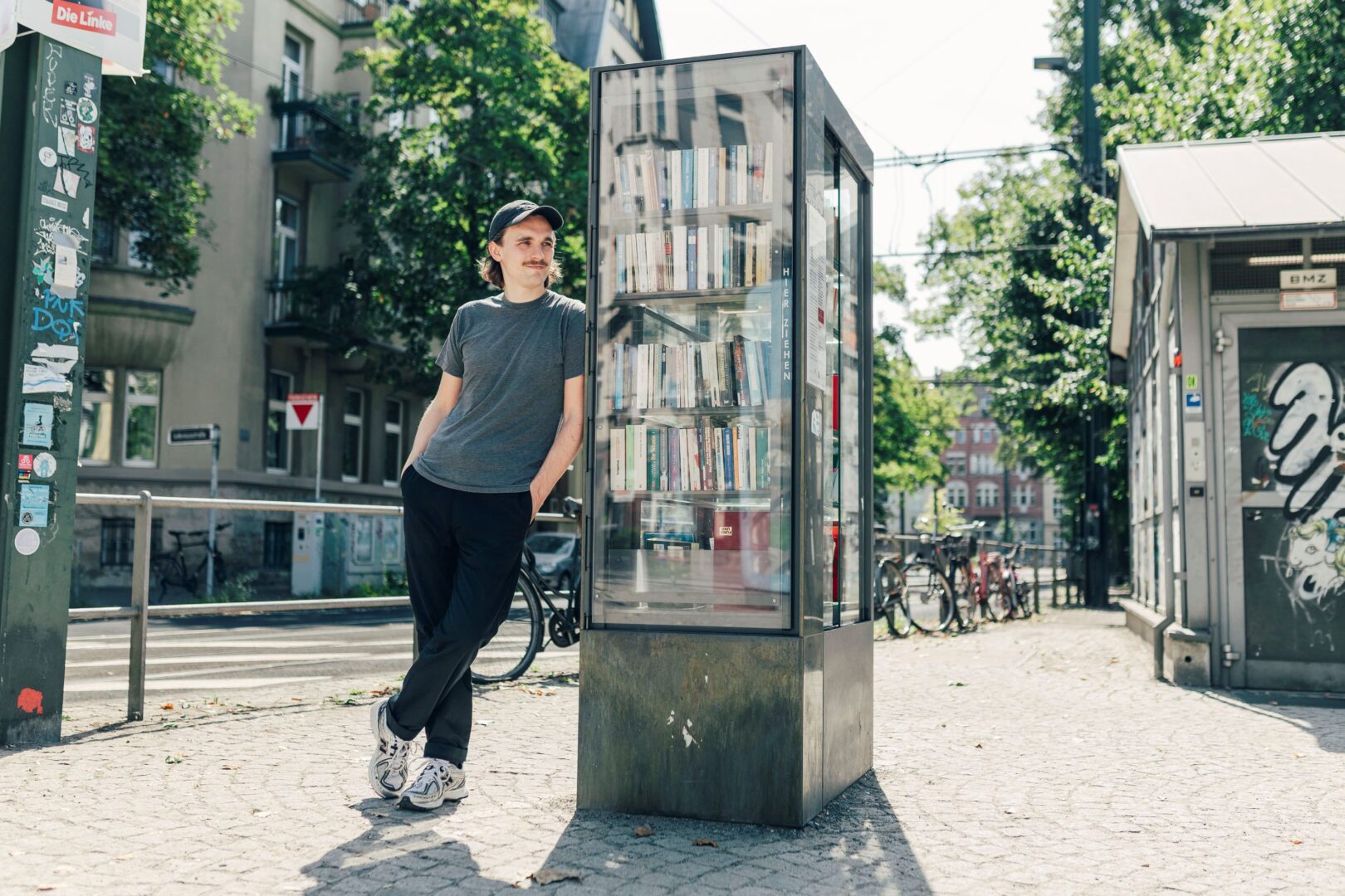 Kaleb Erdmann lehnt sich an einen öffentlichen Bücherschrank. Man sieht im Hintergrund Bäume, Straße und links einen Laternenpfahl, der viele Aufkleber hat.