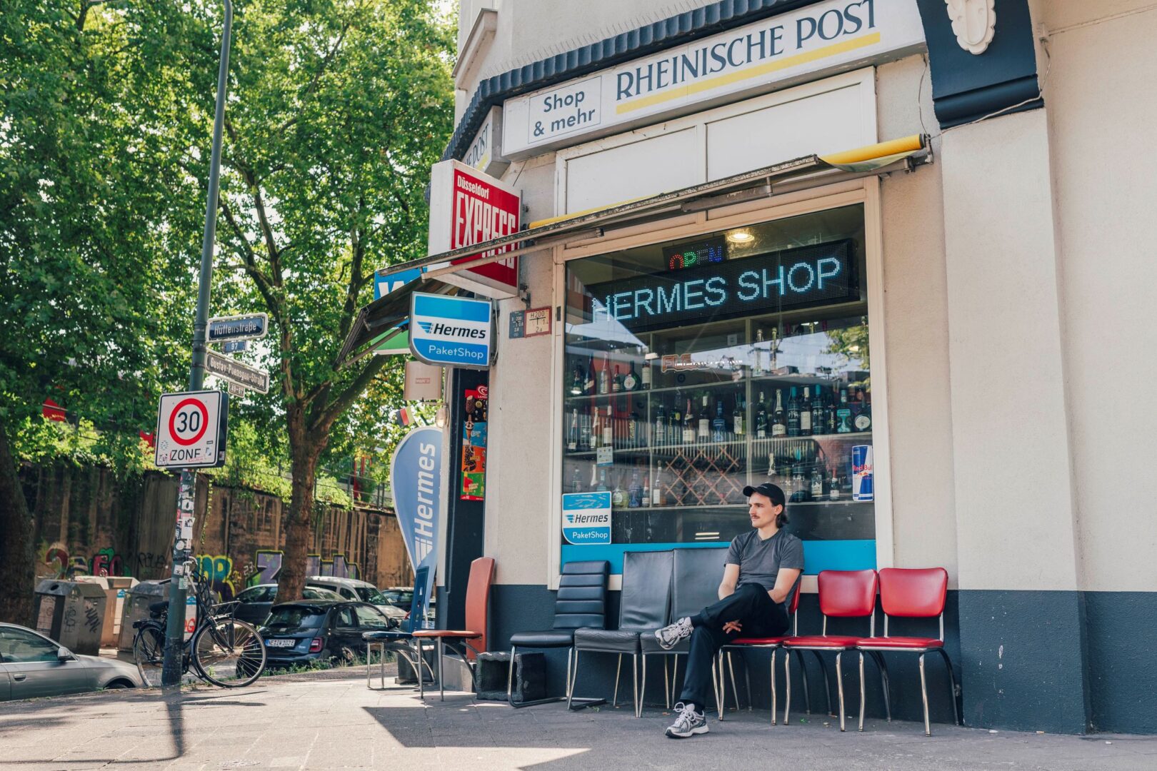 Kaleb Erdmann sitzt auf einem Stuhl vor einem Eckbüdchen in Düsseldorf. Es steht eine Reihe bunt zusammengewürfelter Stühle vor dem Büdchen.